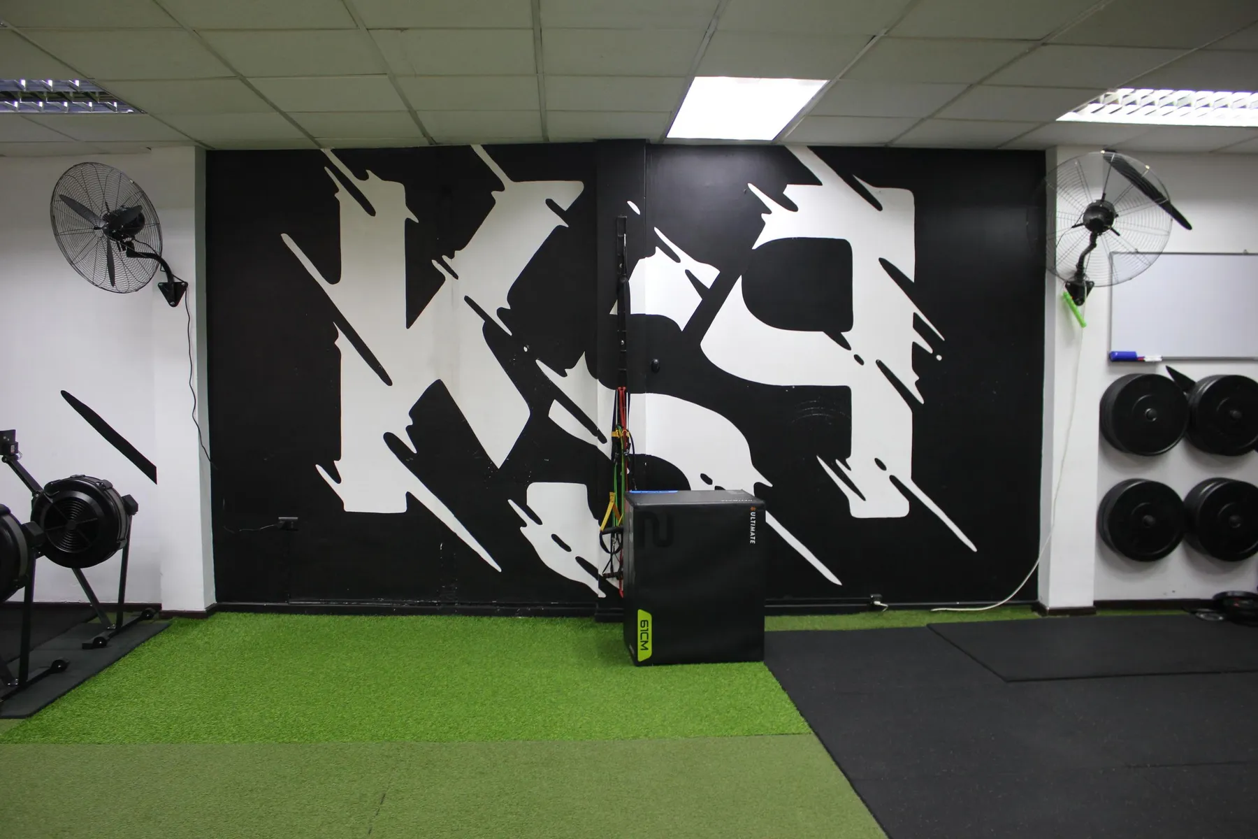 Mural con las letras KSP en blanco sobre muro negro, en la sala de entrenamiento con pasto sintético